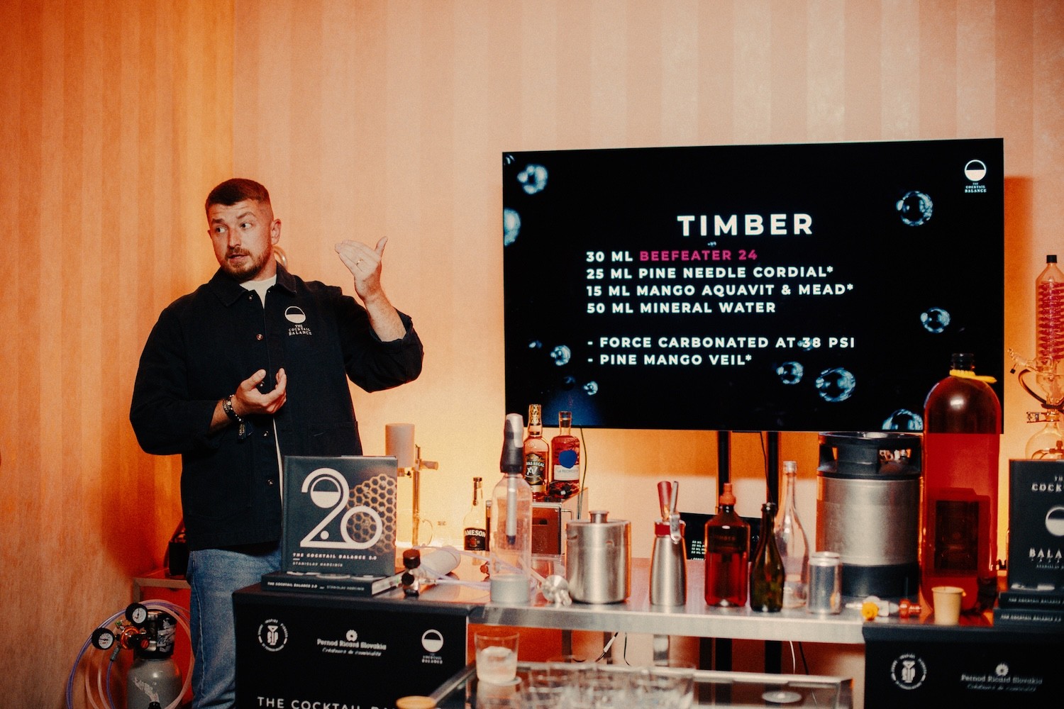 Barman v čiernom polo tričku prezentuje recept na koktail na veľkej obrazovke počas pohostinského podujatia, na stole pred ním sú fľaše destilátov a barmanské náradie, celú scénu zahaľuje teplé oranžové osvetlenie.