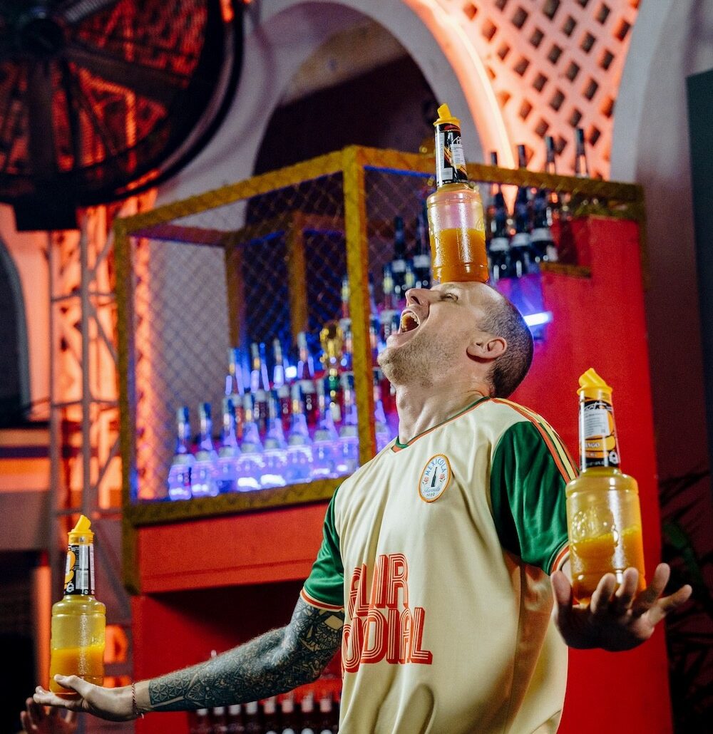 Barman v krémovo-zelenej športovej veste žongluje s fľašami na pódiu počas súťaže vo flair bartendingu, v pozadí neónová kulisa s červeno-oranžovým geometrickým scénografickým dizajnom.