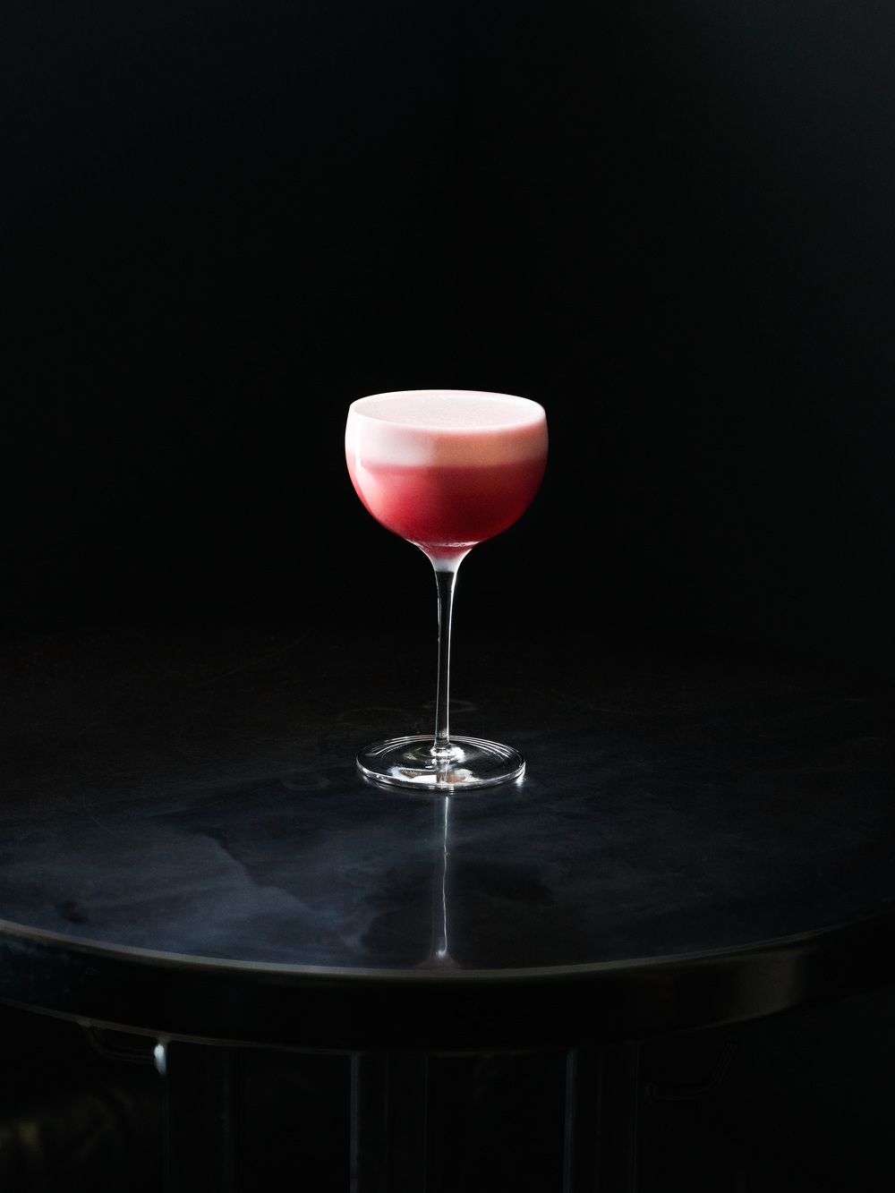 Červený cocktail s bielou penovou vrstvou v coupe pohári na tmavom reflexnom povrchu na čiernom pozadí.