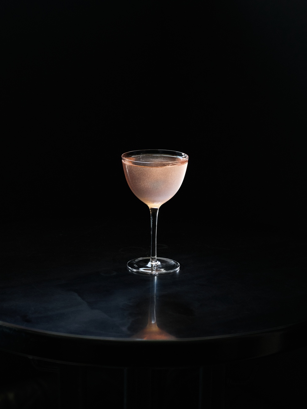 Jediný coupe pohár naplnený bledoružovým cocktailom, stojaci na tmavom reflexnom povrchu na čiernom pozadí s jemným štúdiovým osvetlením.