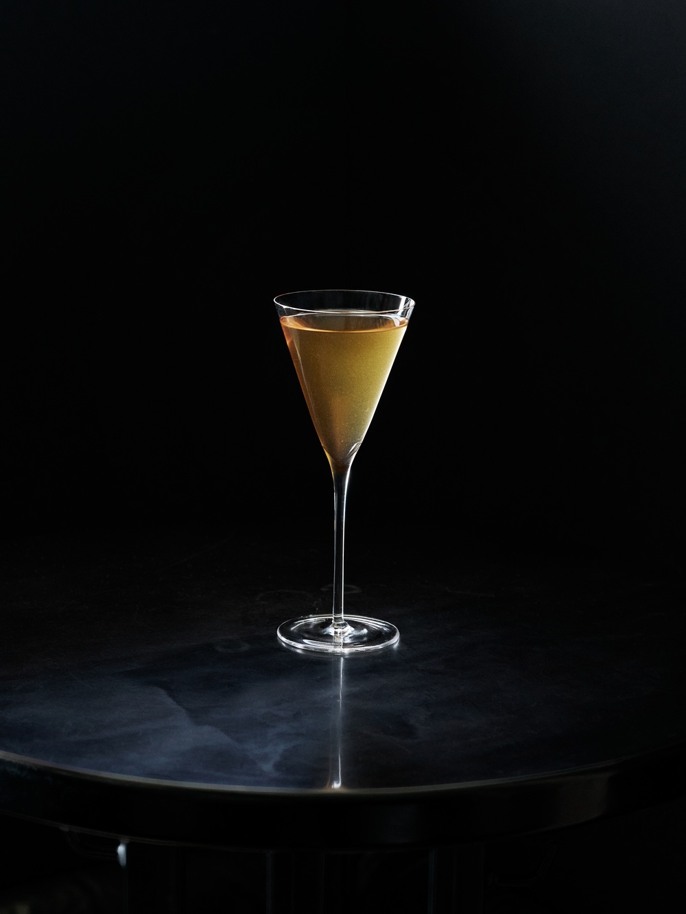 Zlatožltý cocktail v elegantnom coupe pohári na čiernom pozadí, osvetlený dramatickým bočným svetlom vytvárajúcim odraz.