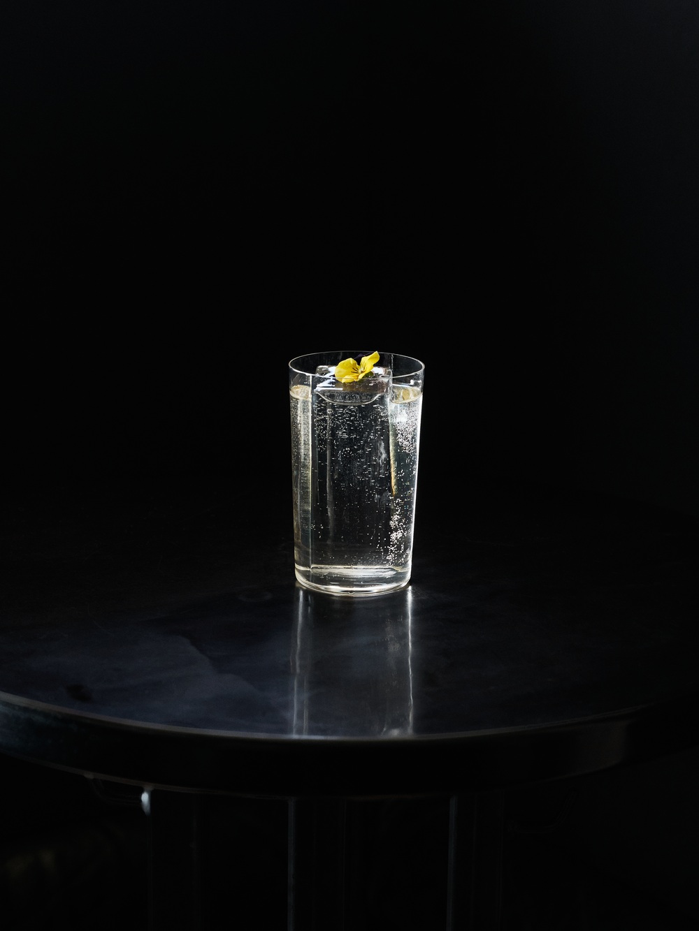 Vysoký highball pohár naplnený perlivou čírou tekutinou s ľadom, ozdobený žiarivou žltou citrónovú kôrkou, fotografovaný na čiernom pozadí na tmavom reflexnom povrchu.