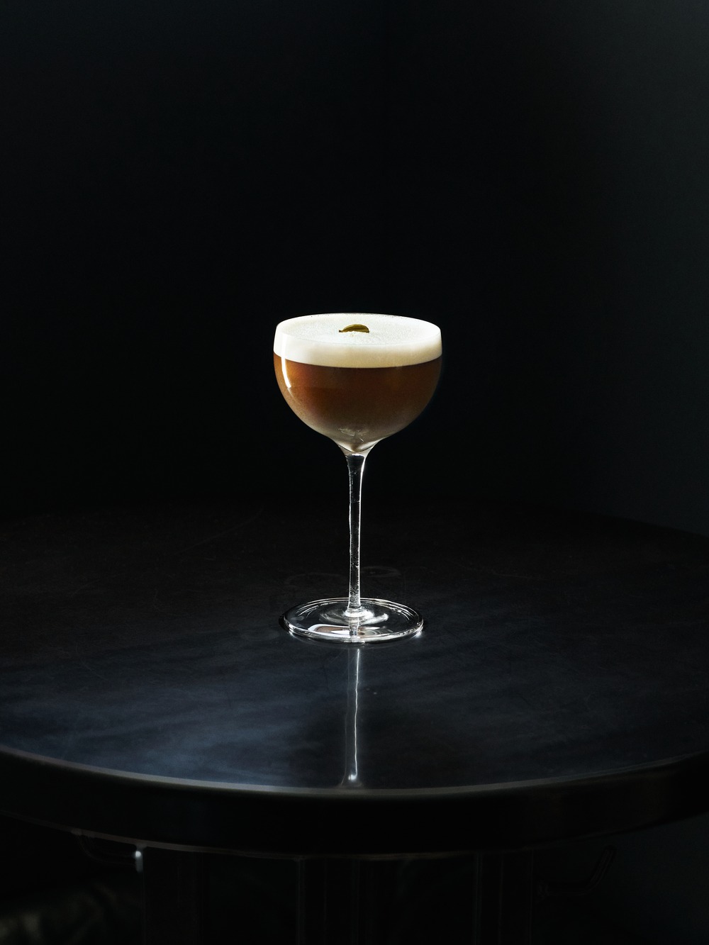 Silueta cocktailu v coupe pohári s hustou krémovou penou a ozdobou z jedného lístka, na tmavom reflexnom povrchu na čiernom pozadí.