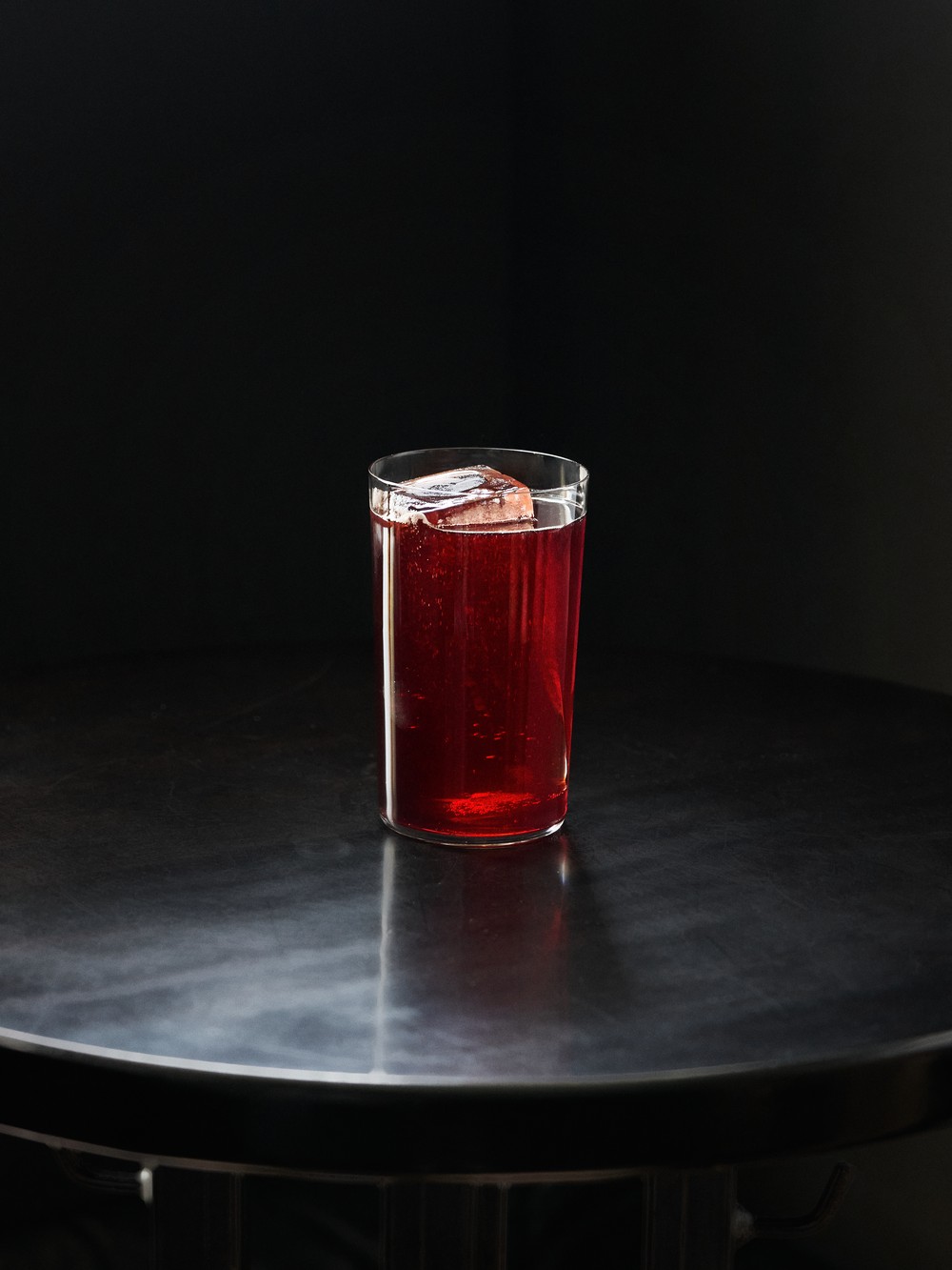 Hlbokočervený cocktail vo vysokom pohári s veľkou kockou ľadu, stojaci na tmavom reflexnom povrchu na čiernom pozadí.