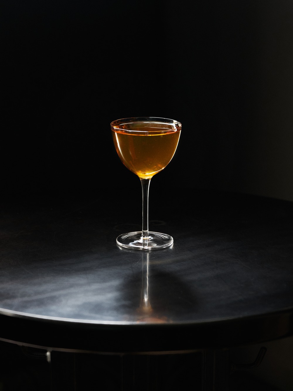 Zlatoambrový cocktail v coupe pohári na čiernom pozadí, stojaci na reflexnom tmavom povrchu s dramatickým osvetlením.