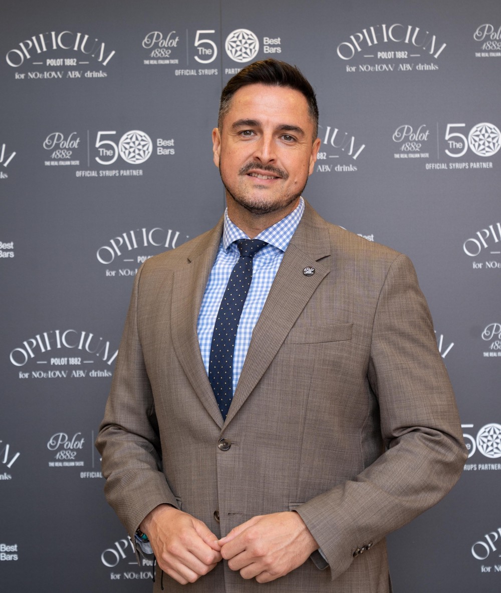 Muž v béžovom saku a svetlomodrej kockovanej košeli s tmavomodrou kravatou na pozadí brandovaného eventového banera s logami Opilici a Polot 1882.