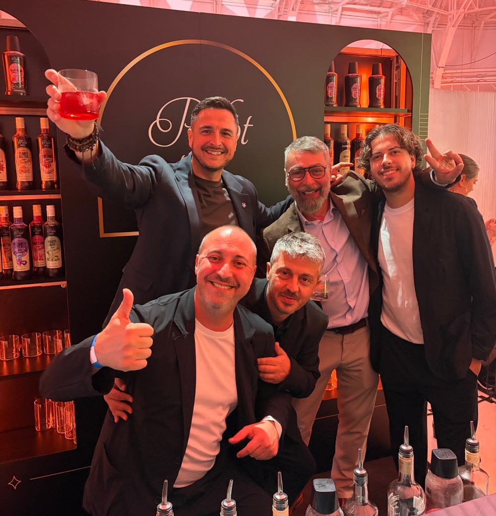 Piati muži v obchodnom oblečení pózujú spolu pred tmavou stenou so žiariacim oranžovým kruhom a neónovým nápisom, pričom dvíhajú červený cocktail. Za nimi sú na poličkách vystavené osvetlené fľaše.